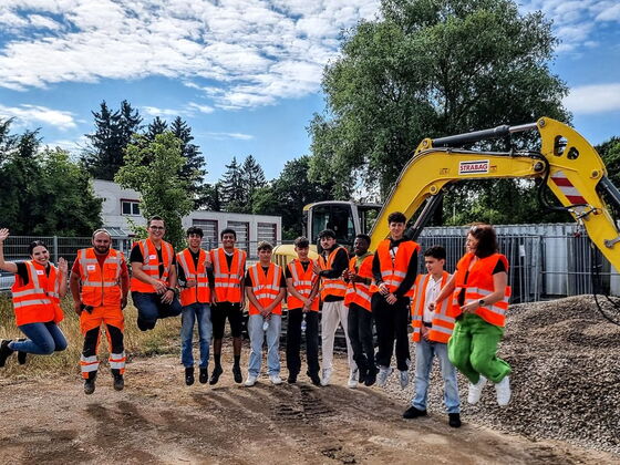 Schüler der 8. Klasse zusammen mit Mitarbeitern hüpfen vor einem Bagger in die Luft